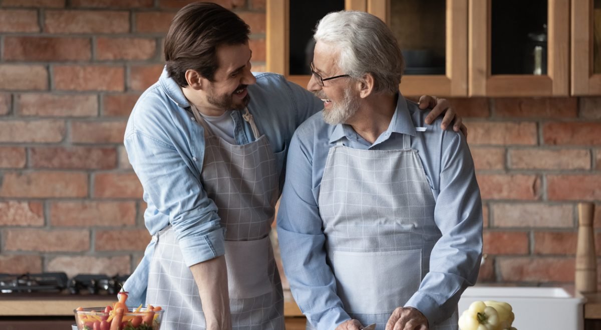 IStock/fizkes - Imagem de Filho amoroso abraçando ombro do pai e cozinhando juntos